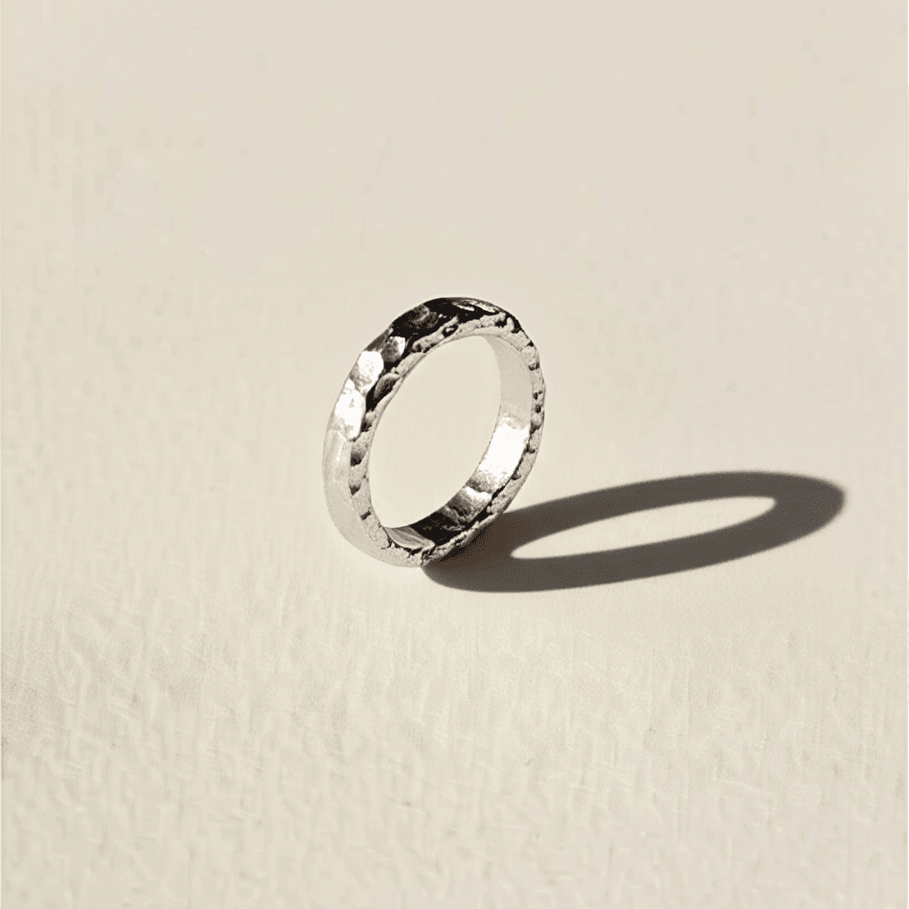 A hammered silver-colored band standing on a warm surface and casting a long shadow.
