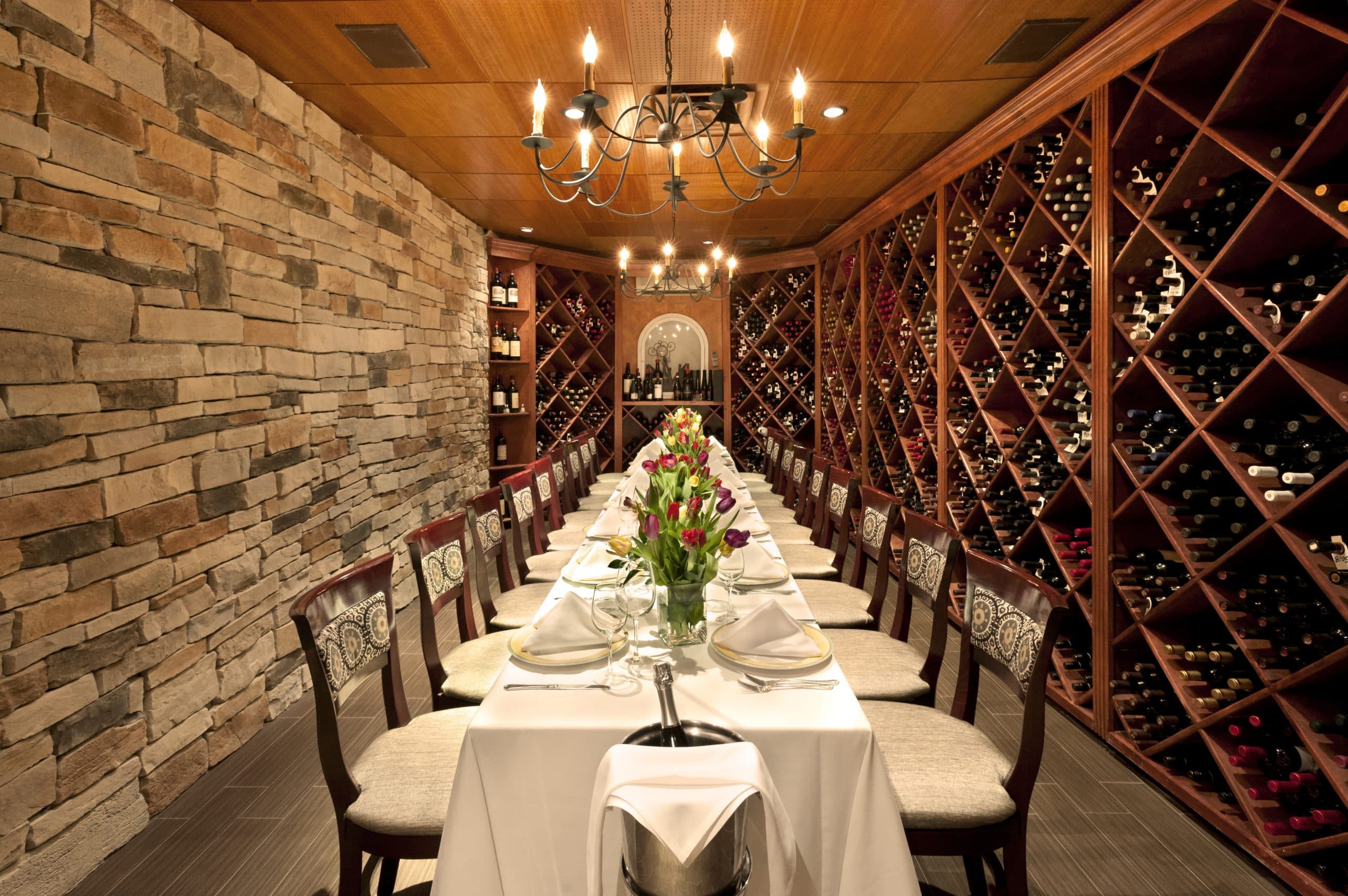 A long dining table set with white linen and flowers in a stone-walled wine cellar.